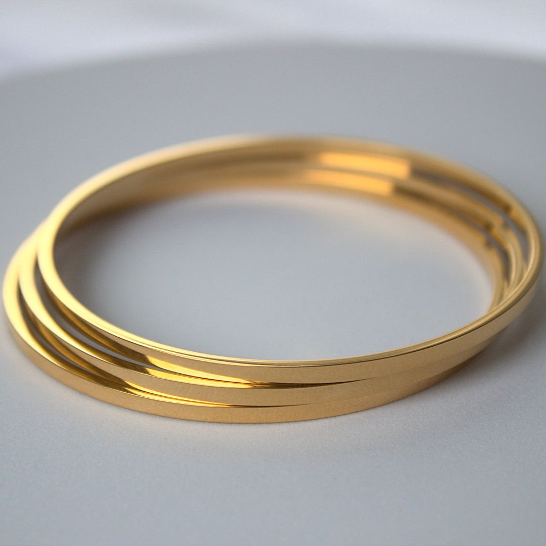 3 simple circle closed bangles in gold, stacked on top of each other slightly unevenly, sitting on white/grey surface.
