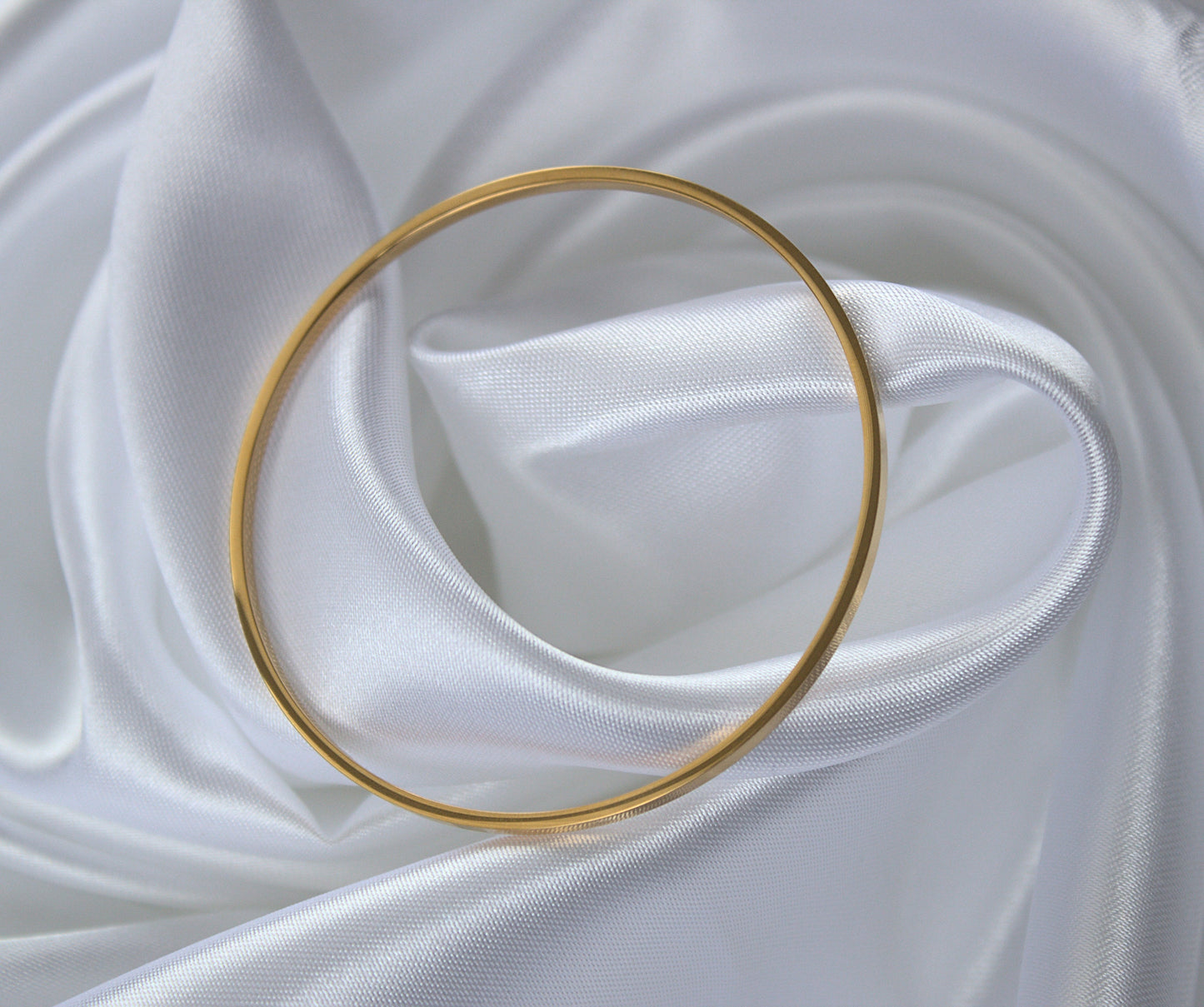 Flat-edged gold circle bangle resting on swirling silk material, top profile.