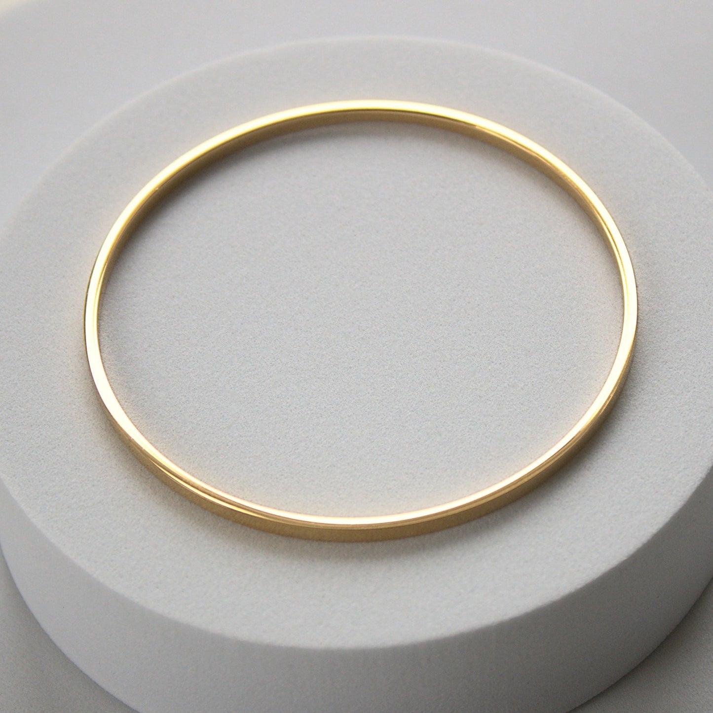 Flat-edged simple gold bangle resting on white cylinder surface. Top profile showing whole bangle with light shining on the flat edge.