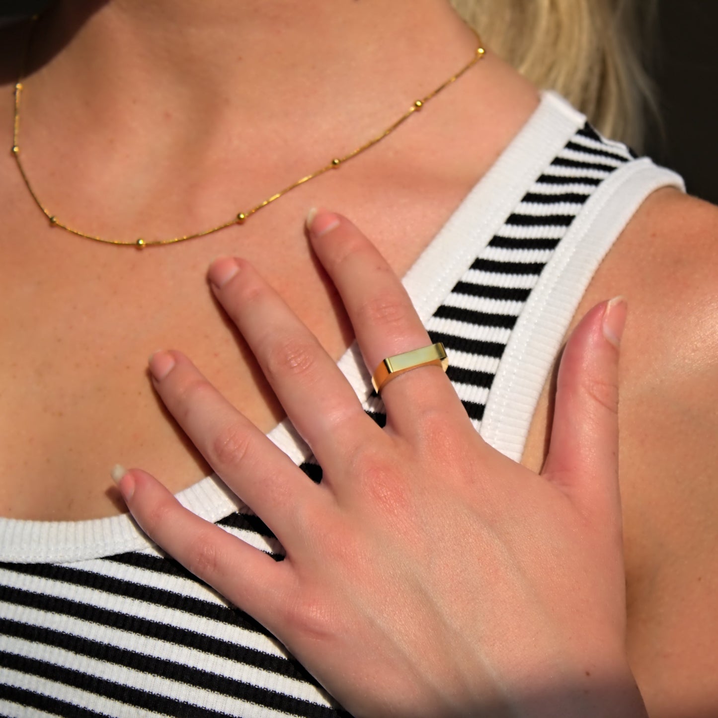 Gold flat bar signet ring on model, with hand out-stretched and pressed against top of chest, highlighting flat edge of ring that is facing the camera.