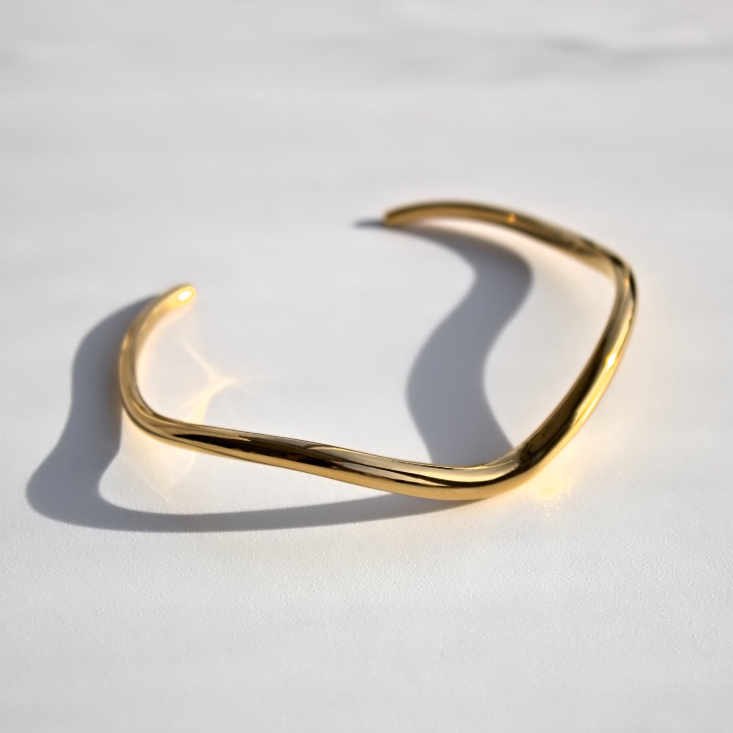 Frontal view of irregular gold cuff bangle with a flowing V shape design. Sun light shining from right side, sitting on plain white surface.