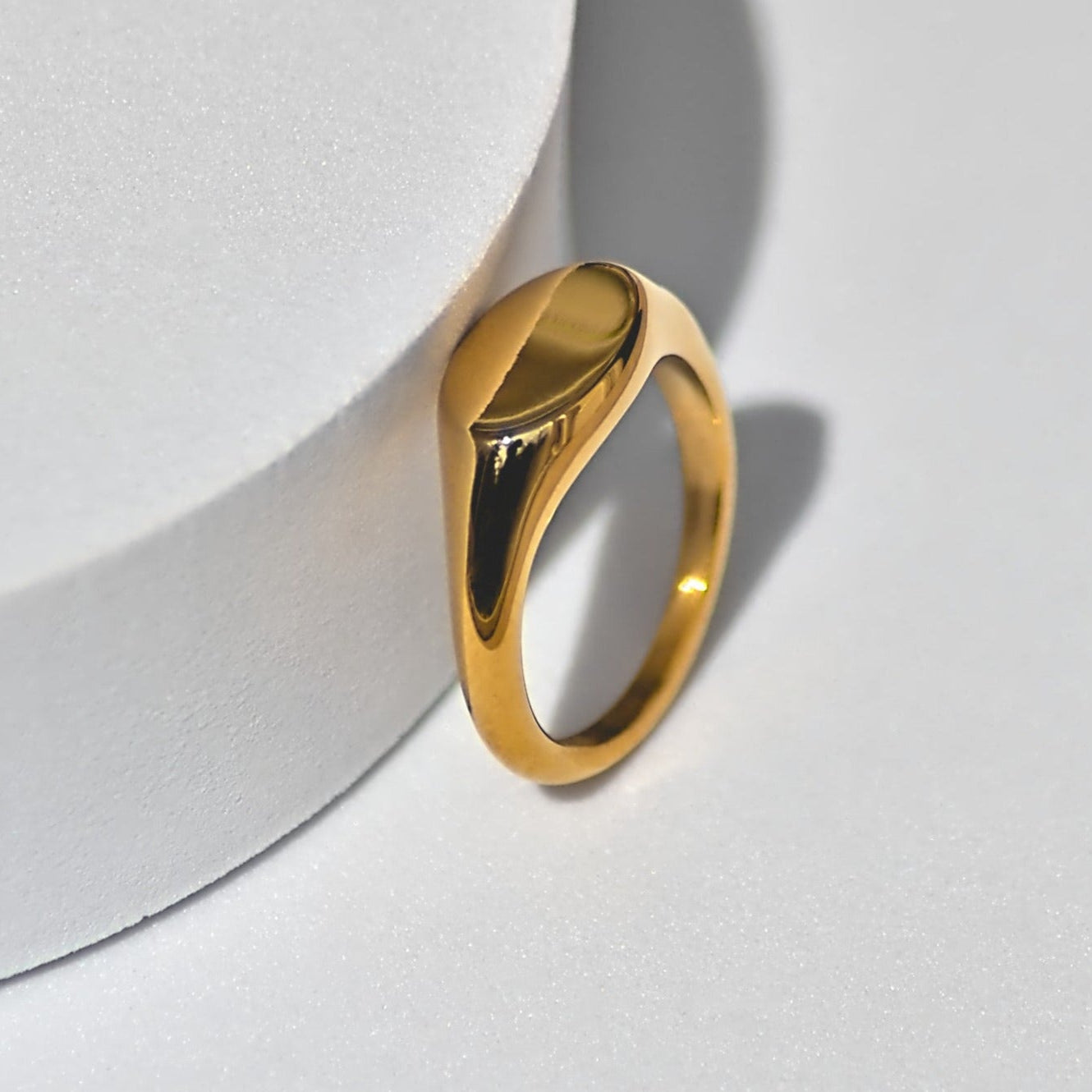 Elegant dainty oval-edged gold ring, leaning against circle shaped white surface, and standing on white surface.