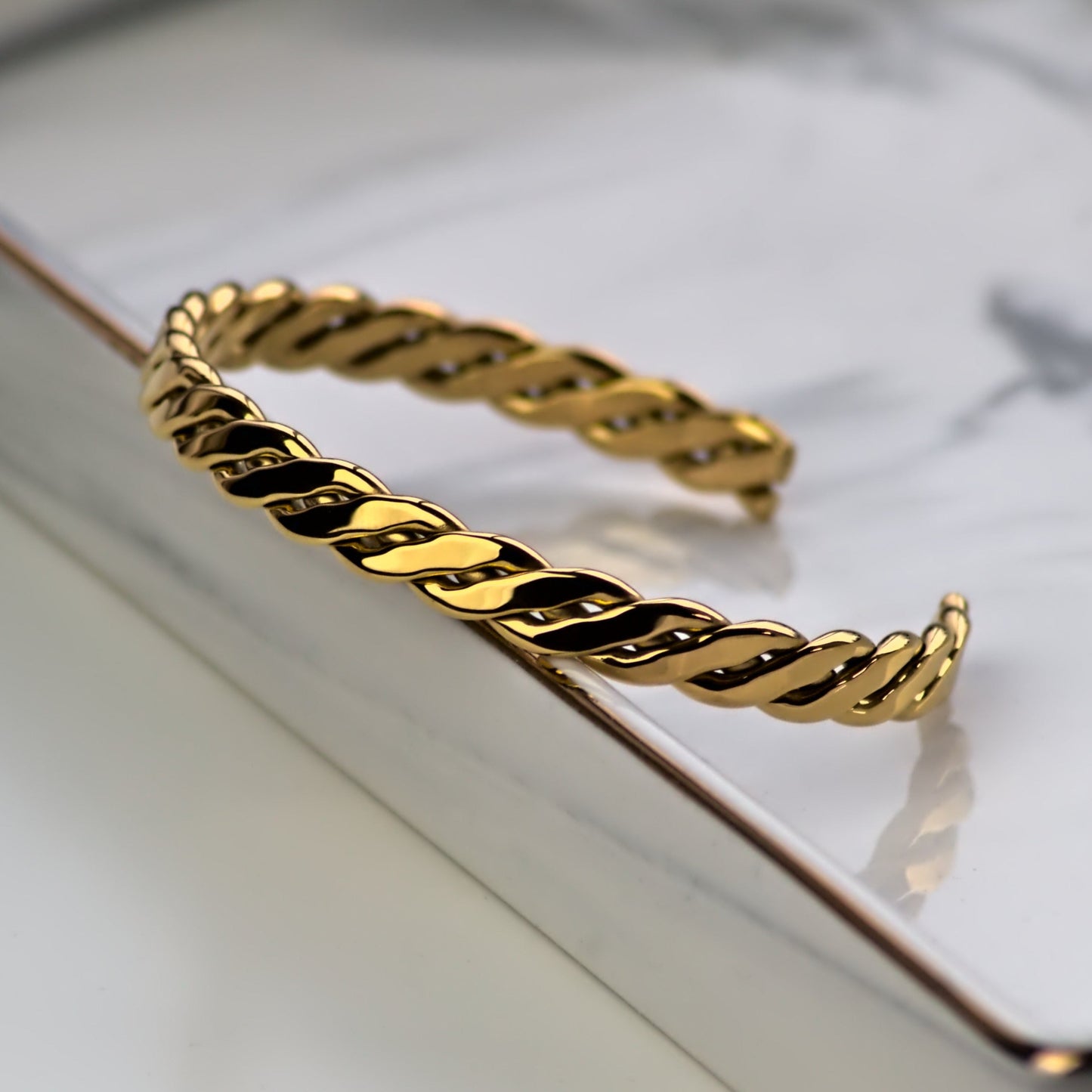 Interweaving and woven style cuff bangle in gold colour, resting against edge of silver square marble tray. Close up on front side of bangle with light highlighting smooth surface of bangle.