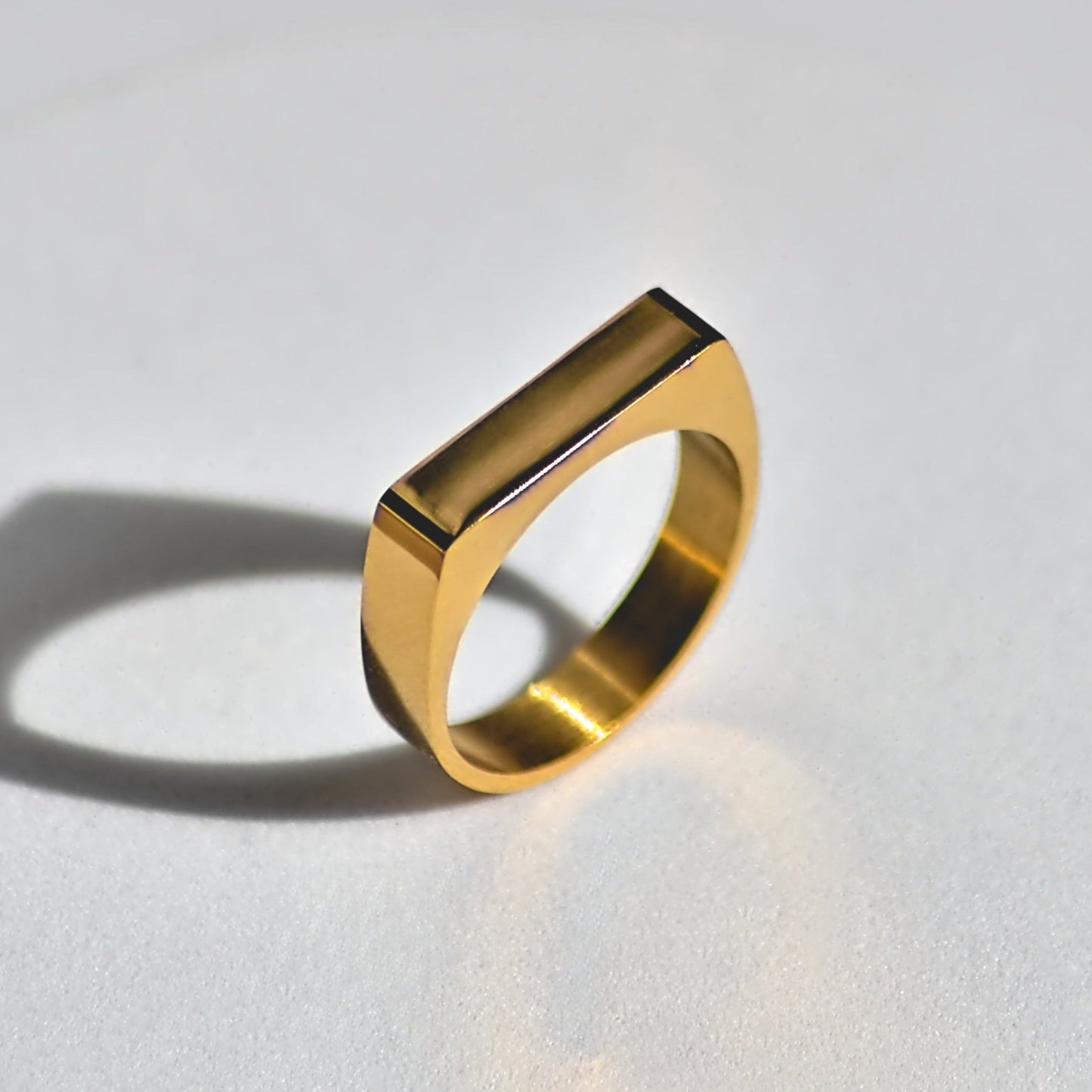 Gold signet ring with square and flat-edged top, balancing upright on white surface at 45 degree angle, and sunlight coming from the right side and casting a long shadow to the left.