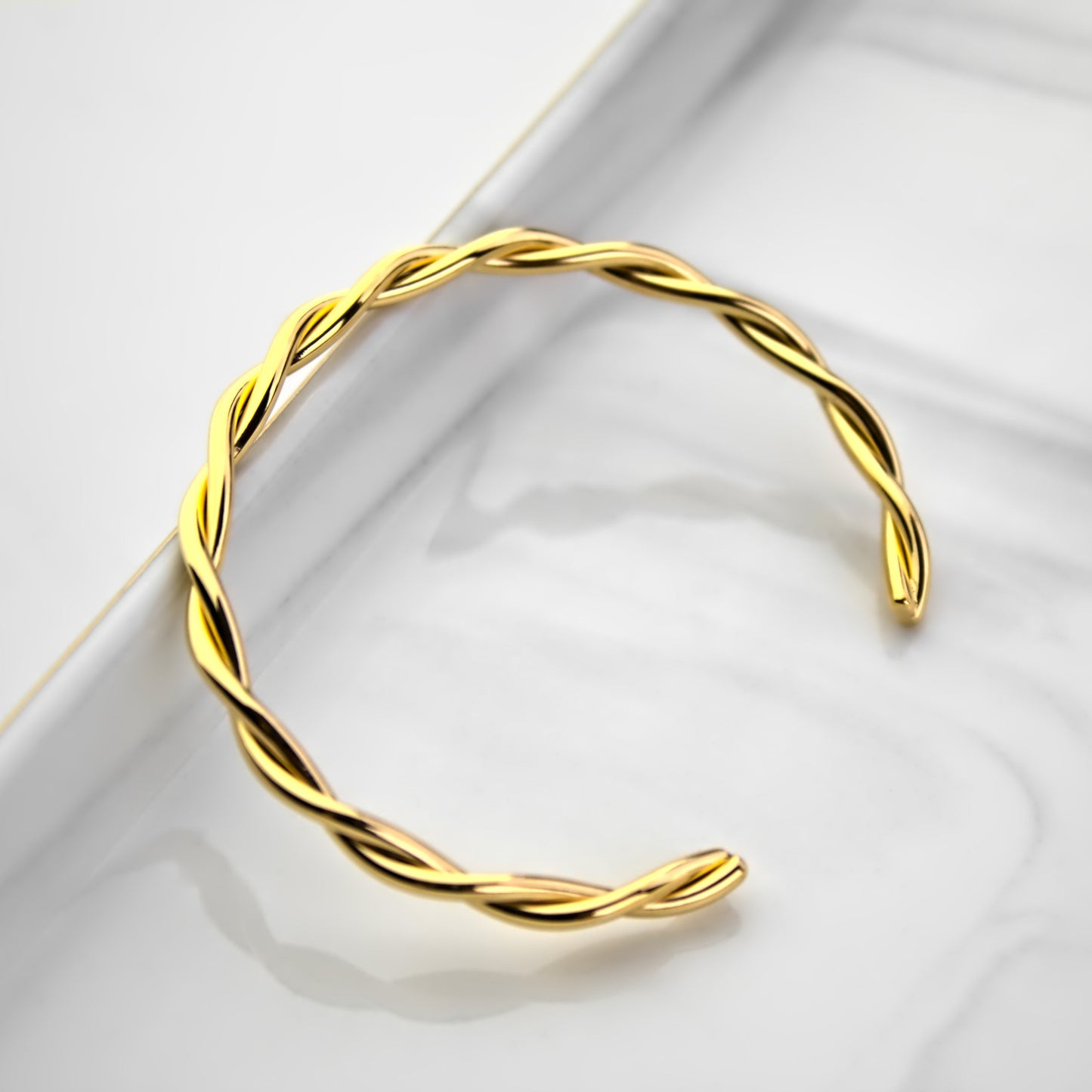 Interweaving gold cuff bangle resting on edge of marble tray, top down profile view with bangle facing sideways.