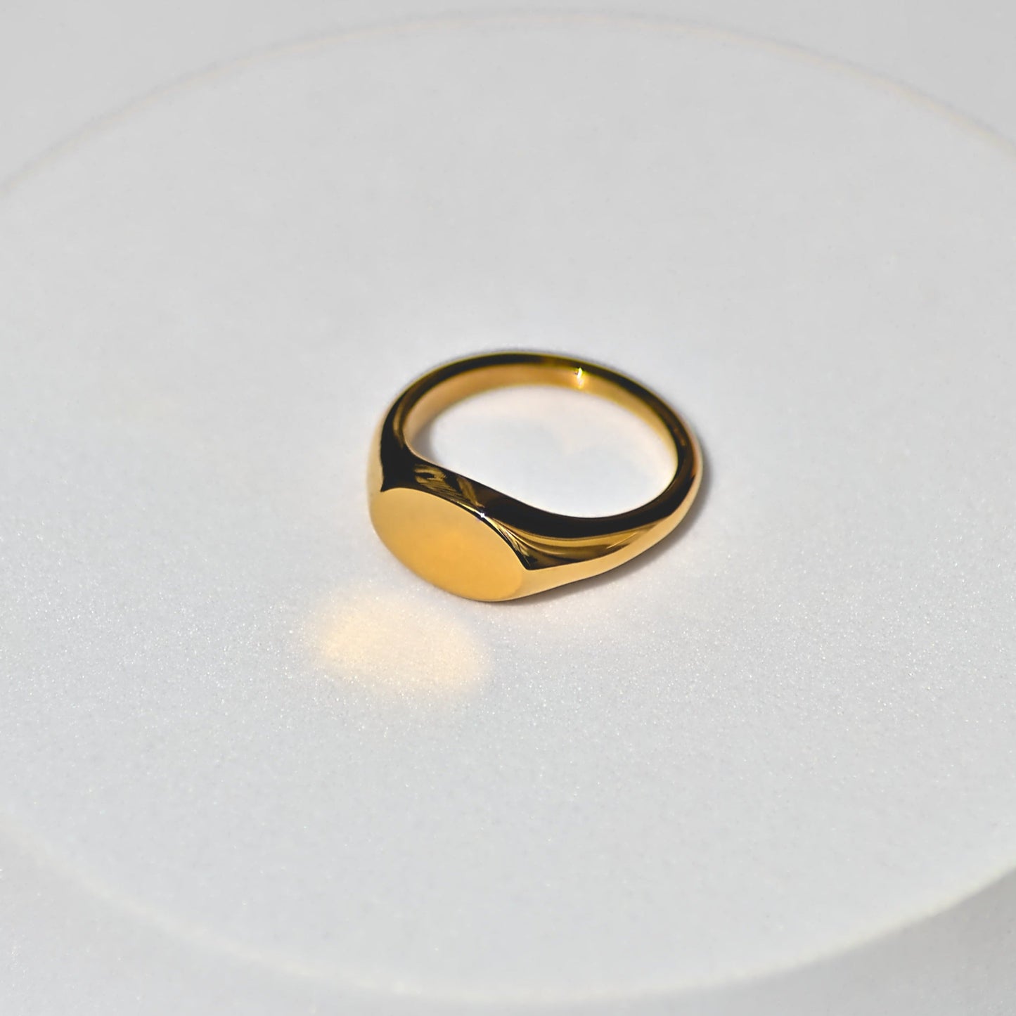 Oval shaped gold signet ring sitting on plain white surface, facing forwards camera slightly leftwards at 7 o clock position.