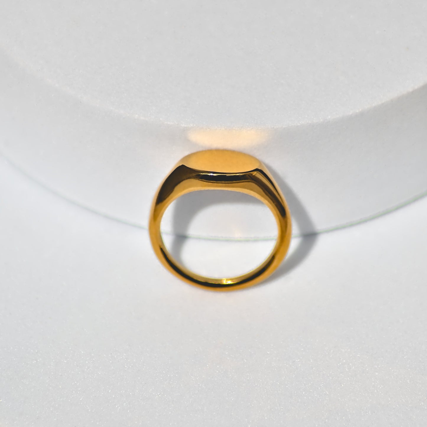Gold oval shaped signet ring on flat white surface, balancing upwards and leaning backwards against white cylindrical shape behind it. Oval feature pointing upwards.