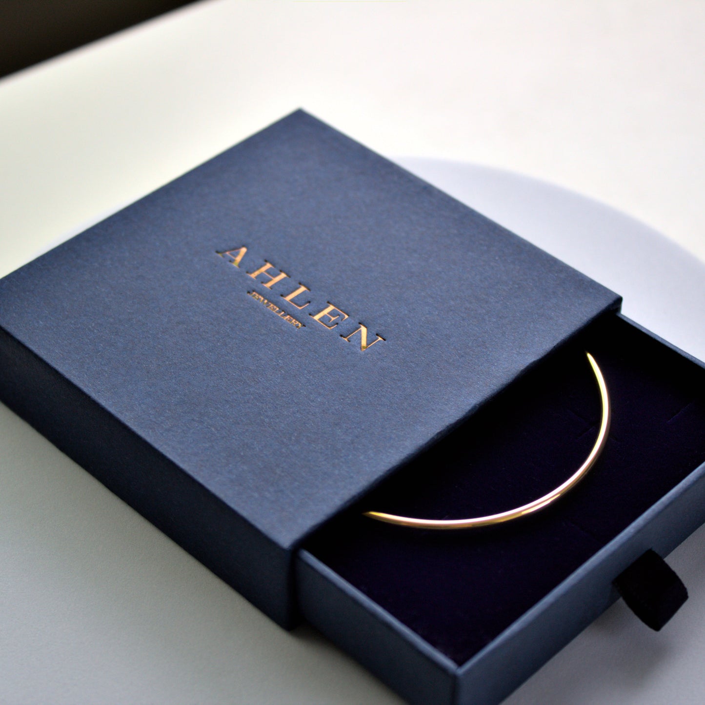 Rounded edge gold circle bangle sitting in Ahlen Jewellery branded blue gift box. Slide out box slightly open, with part of bangle showing inside the box. Debossed gold logo on the box, and box is sitting on white background.