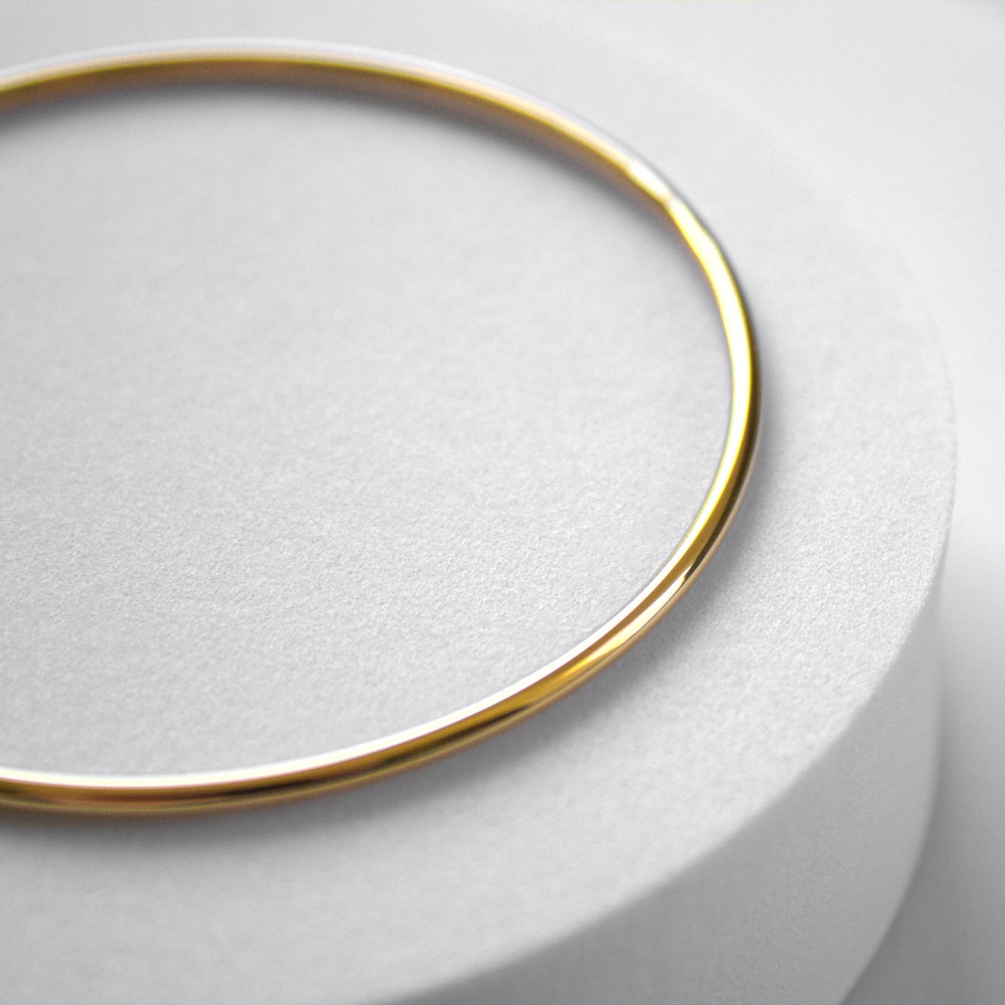 Rounded edge simple circle gold bangle, resting on white cylinder surface. Slight close up, highlighting the rounded shape of the bangle.