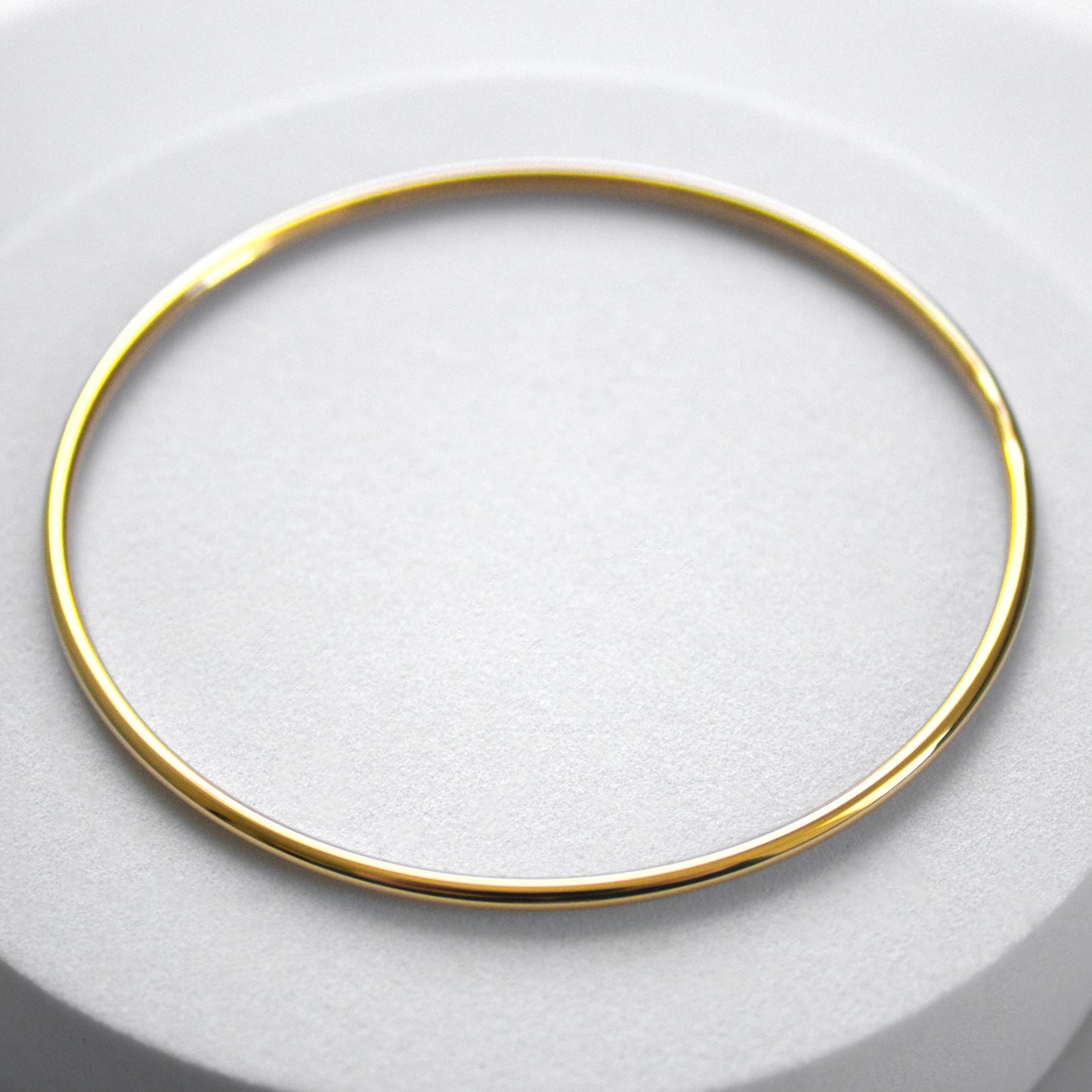 Rounded edge simple circle gold bangle on rounded white surface. Whole bangle in image, top profile.