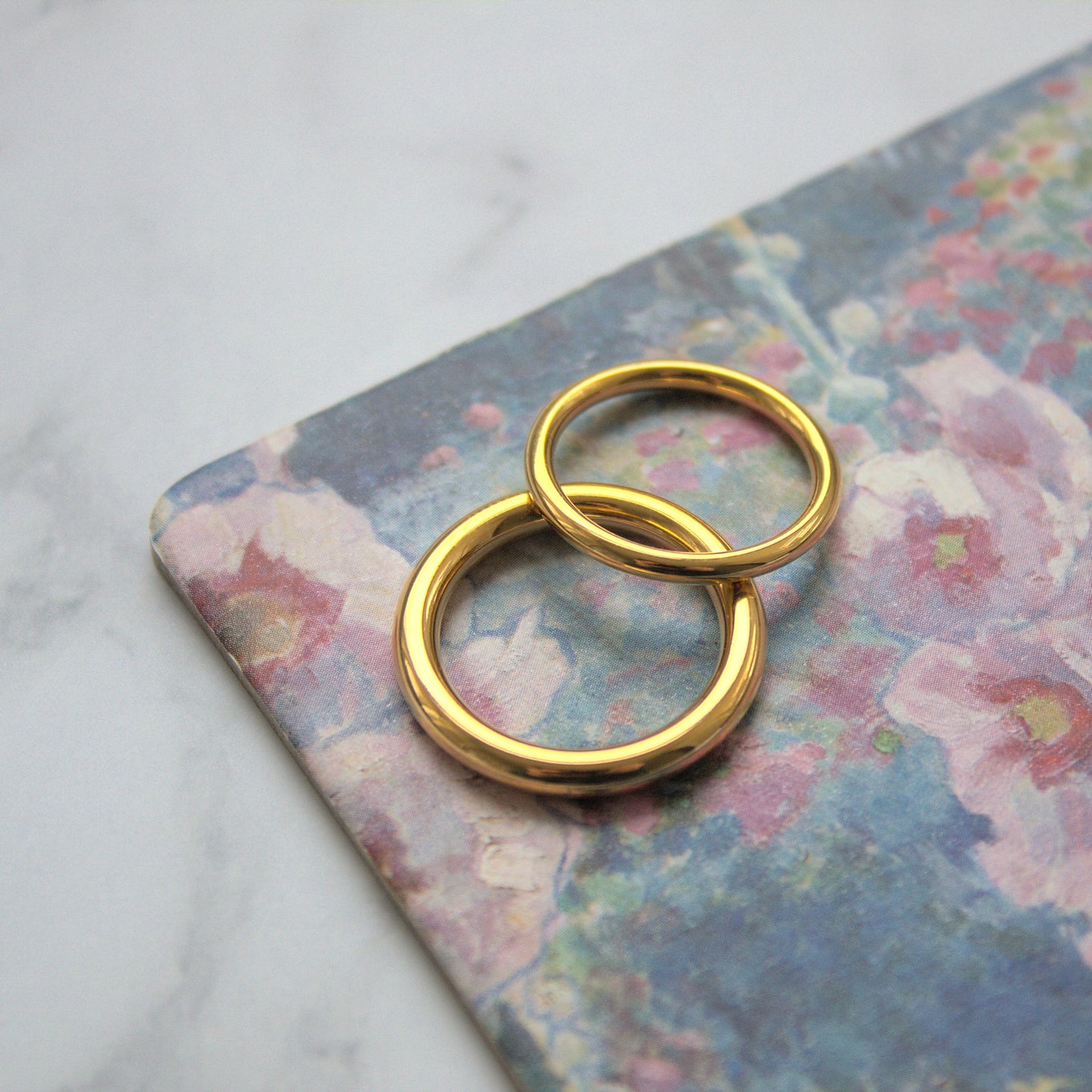 Two simple and elegant circle rings, one thicker and one thinner, resting on flower patterned coaster near corner. Thicker ring flat on coaster, thinner ring slightly above and leaning against the thicker ring.