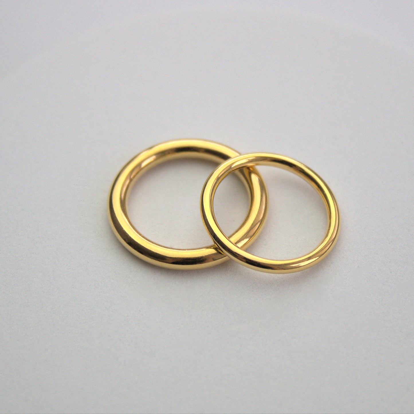 Elegant gold thicker and thinner circle rings. Thicker ring flat down on white surface, thinner ring to the right and leaning on the thicker ring. Smooth polishe finish to the rings.