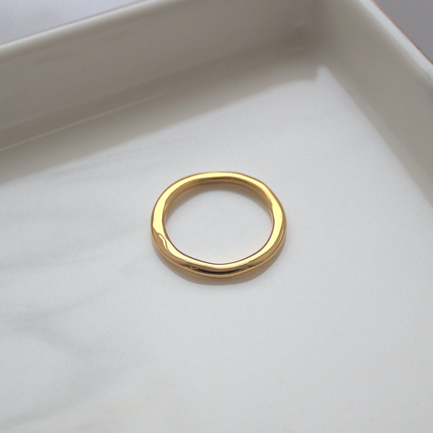 Elegant thinner gold wave ring resting on silver marble tray near corner, light highlighting the smooth ring surface