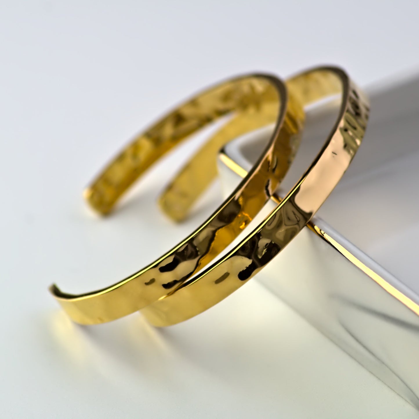 Two gold cuff bangles with hammered finish, both leaning against outer edge of elegant marble tray, with the outer bangle leaning on the other bangle.