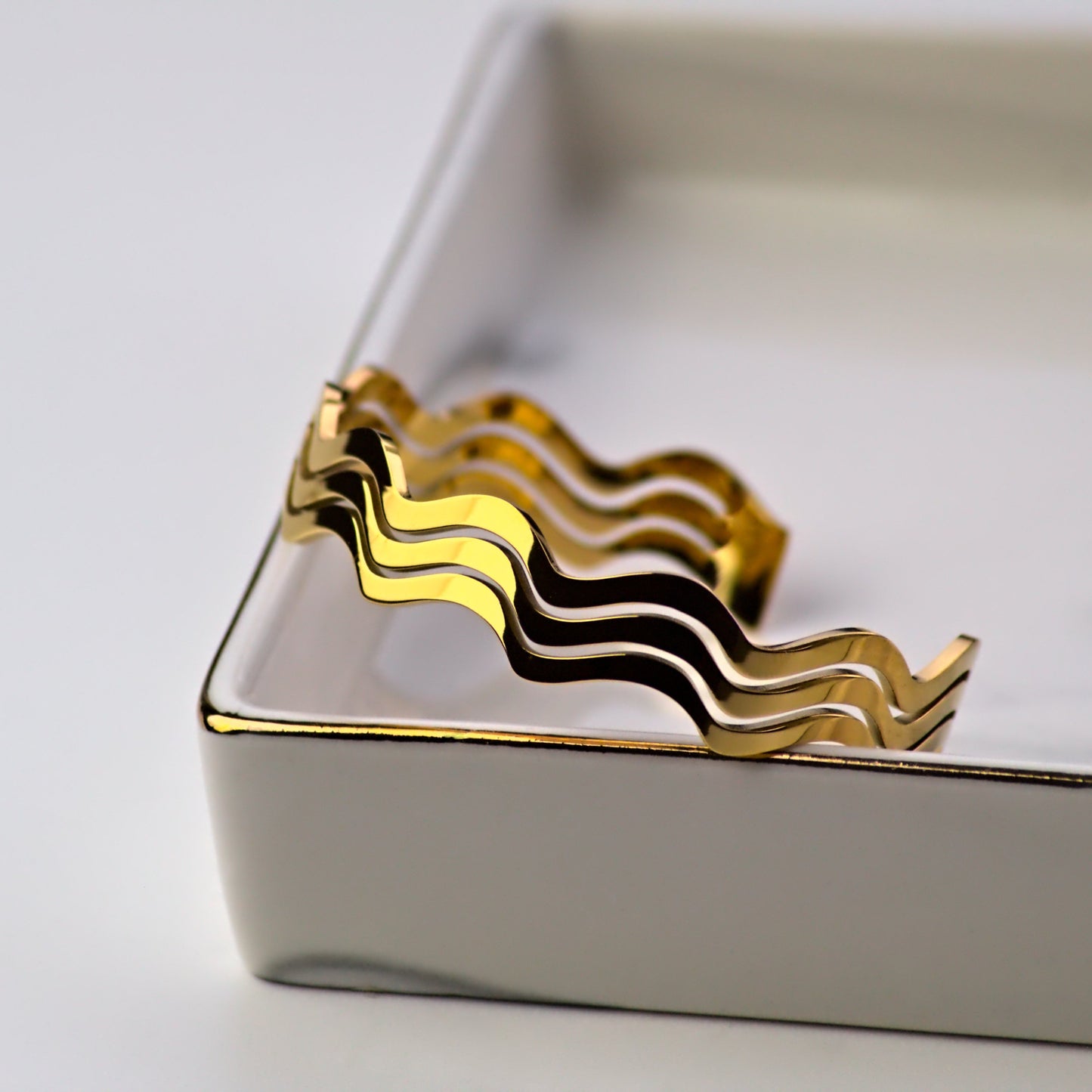 Wavy gold bangle elegantly resting facing outwards from inside of tray, leaning on corner. Light reflecting off surface of bangle.