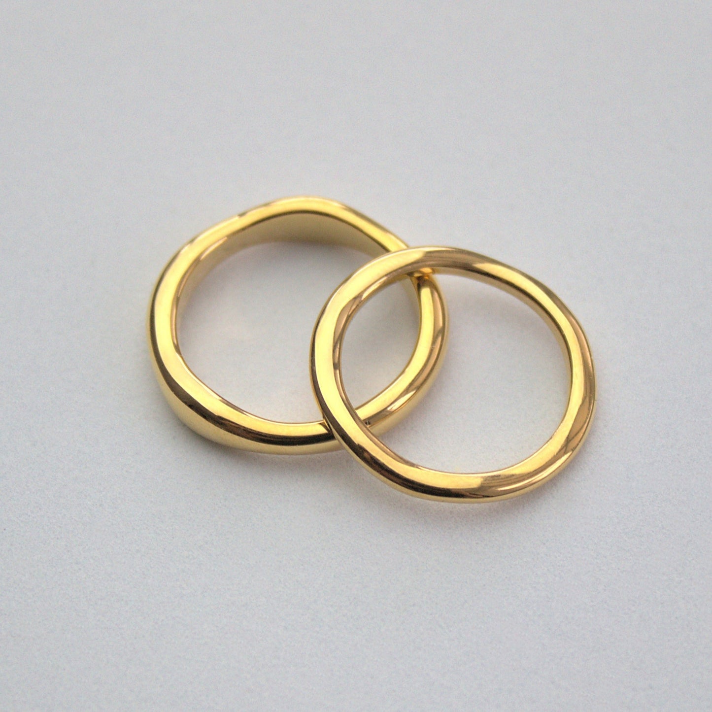 2 elegant gold rings, one sightly thicker on the left, with slightly uneven pattern, thinner ring resting on the side of the thicker ring, and both sitting on a plain white surface.