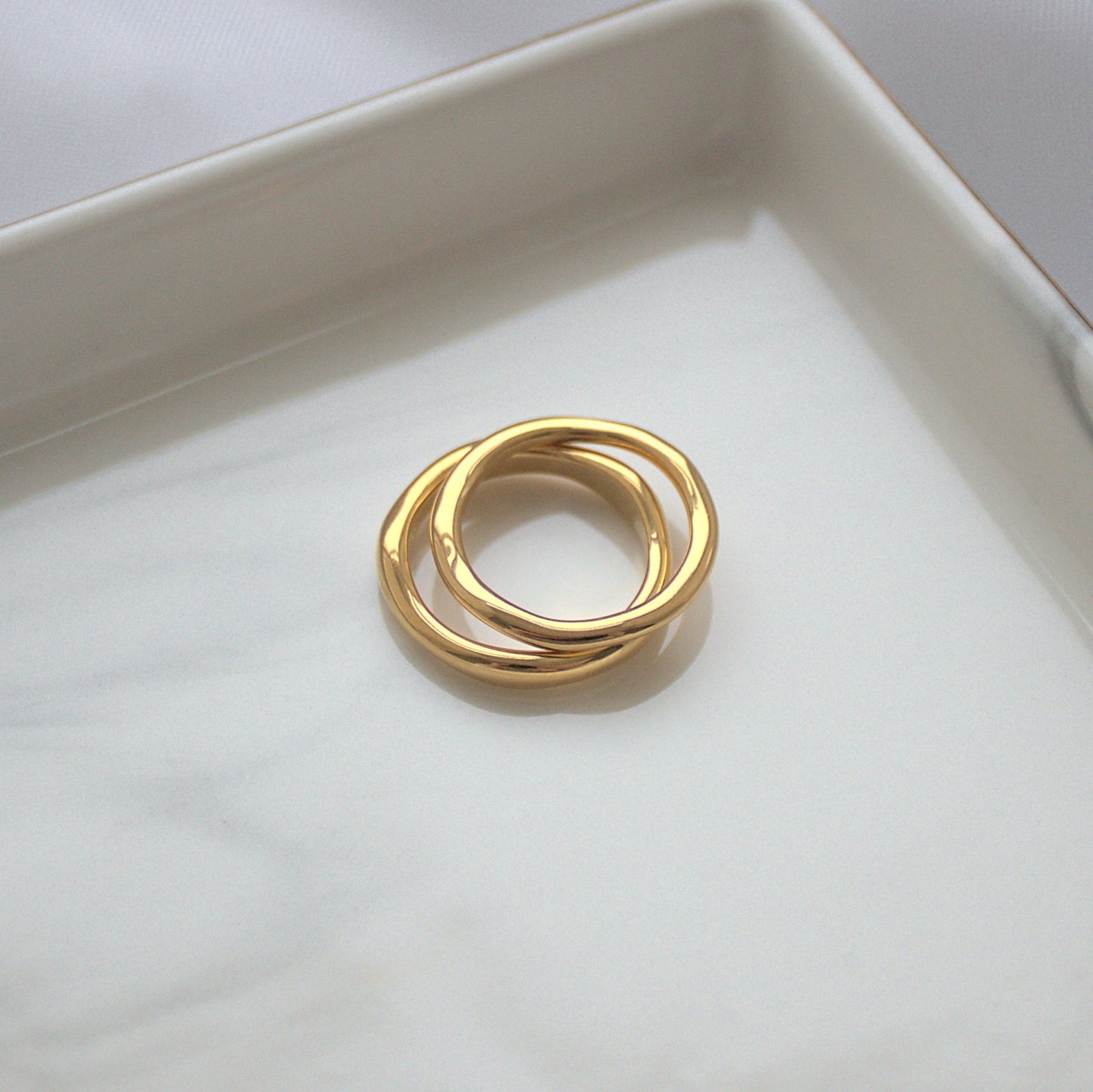 Elegant gold ring set, thinner ring stacked on top of thick ring, sitting on silver marble tray near corner.
