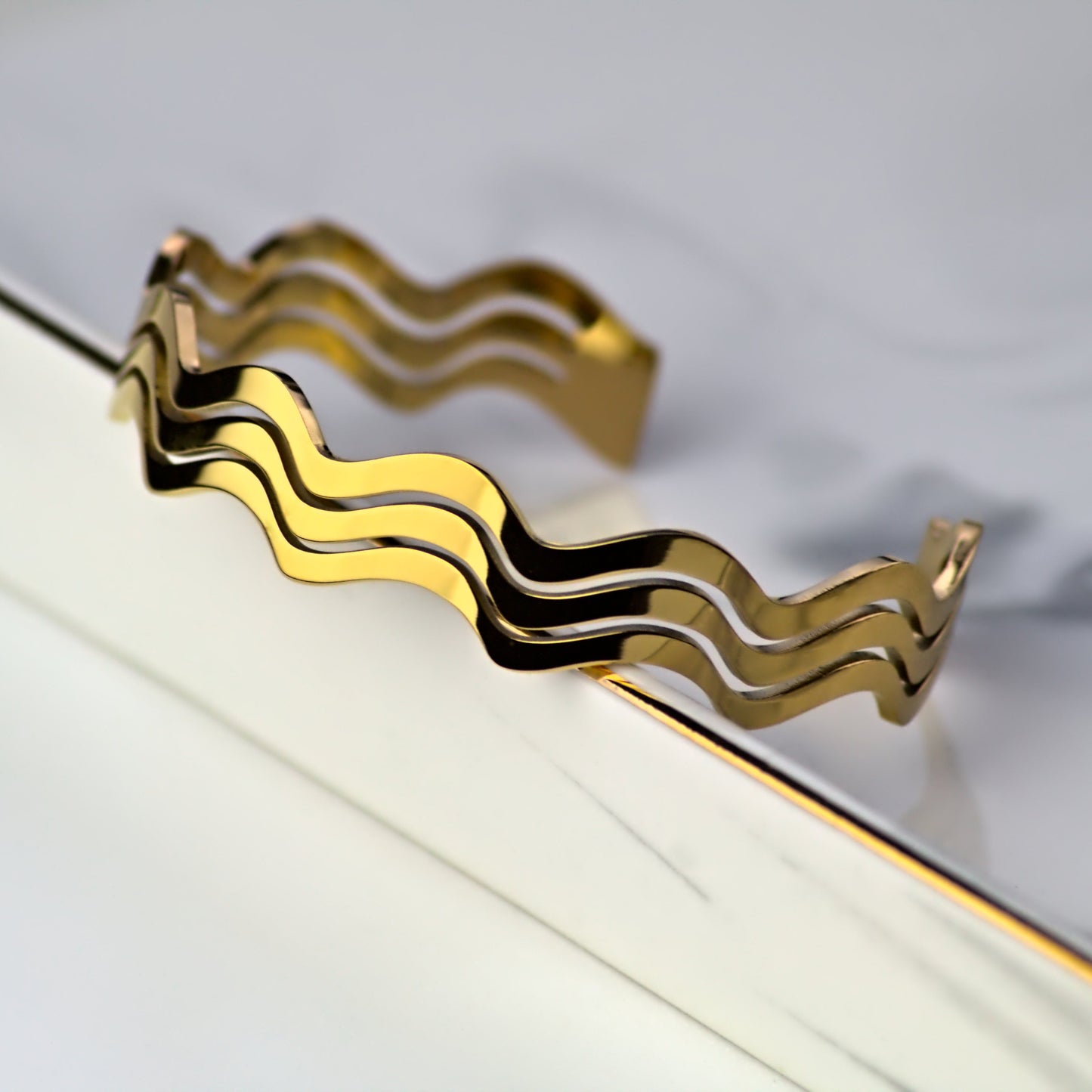 Wavy design cuff bangle with 3 ripple segments, resting on inside edge of square tray, facing outwards. Light reflecting and highlighting smooth surface edge of bangle.