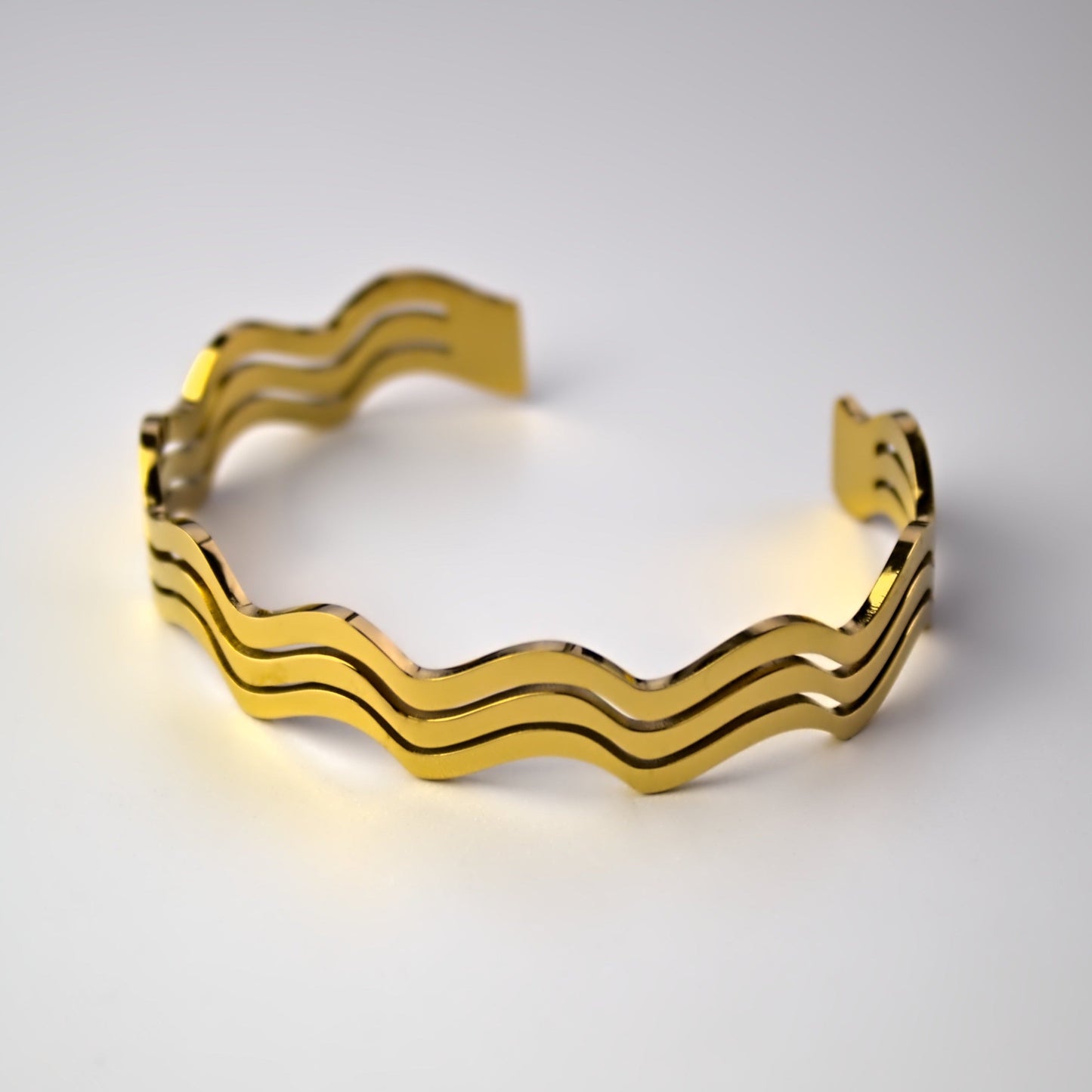 Gold wavy designed cuff bangle resting on flat white surface, front profile.