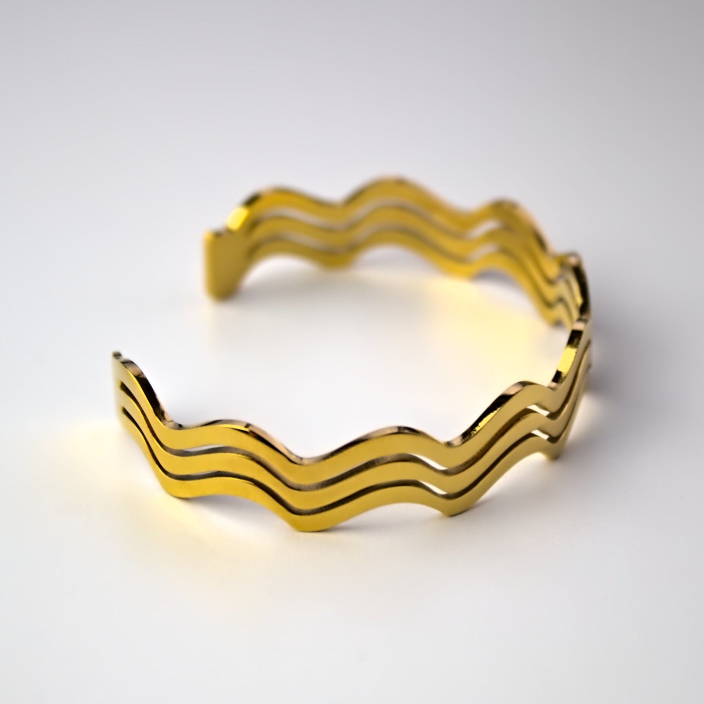 Wavy designed gold bangle resting on flat white surface, sideways view.