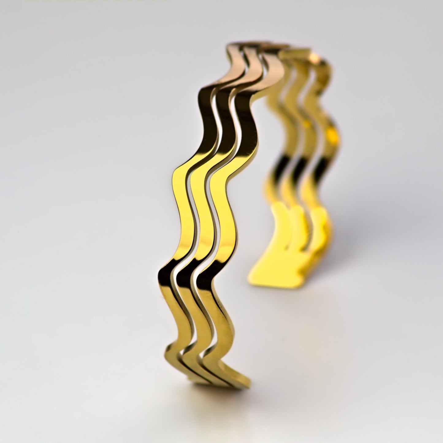 Wave gold bangle standing balanced upright on flat white surface, light reflecting off surface edge.