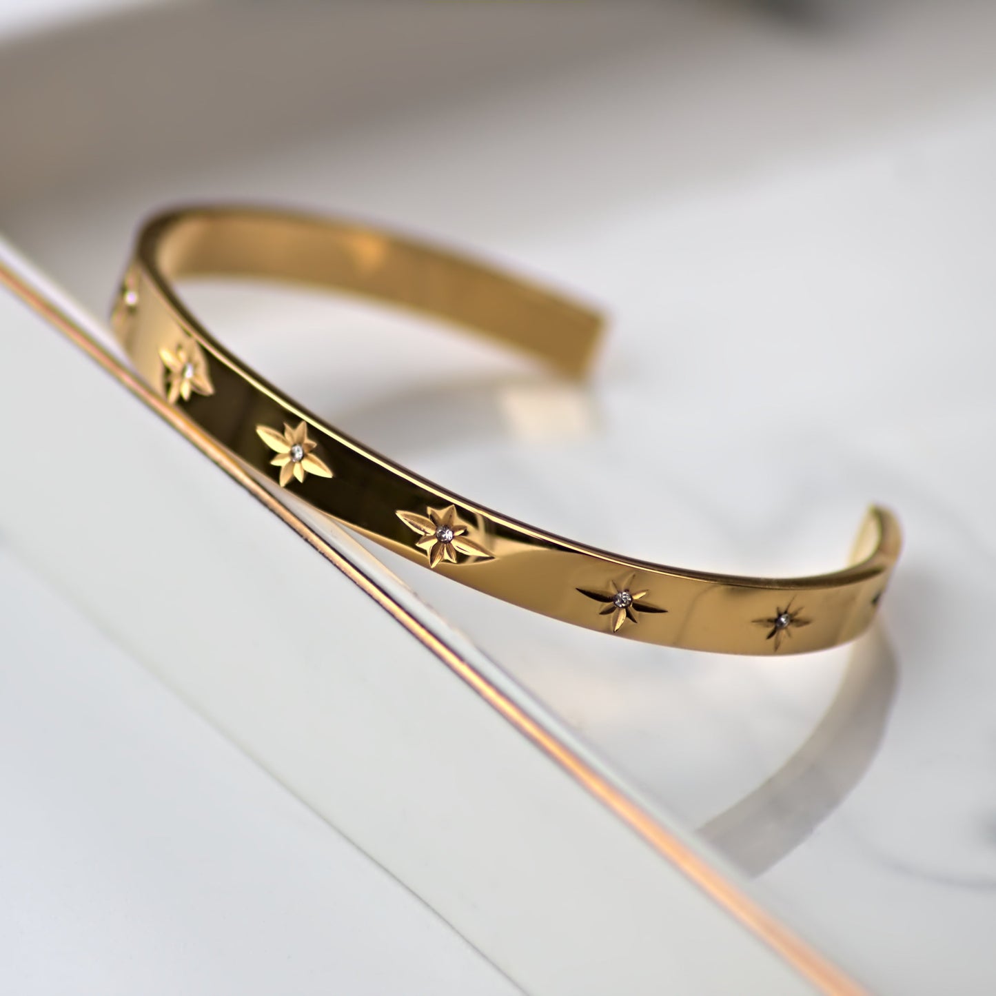 Flat edged cuff bangle featuring zircon stones embedded in star shape pattern, all along the bangle front. Leaning on edge of marble tray.
