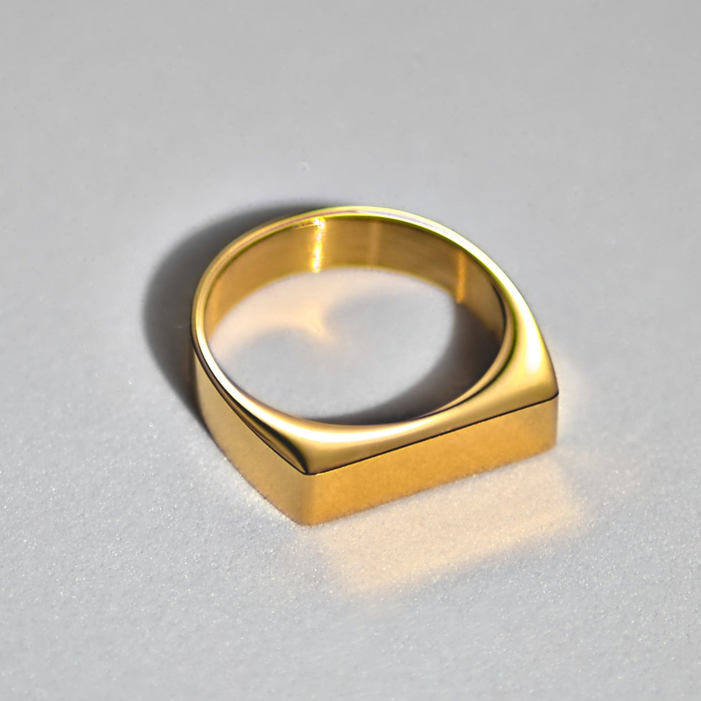 Flat-edged gold signet ring sitting flat on white surface, with strong light reflecting off the top edges. Ring facing forwards at 5 o clock position.