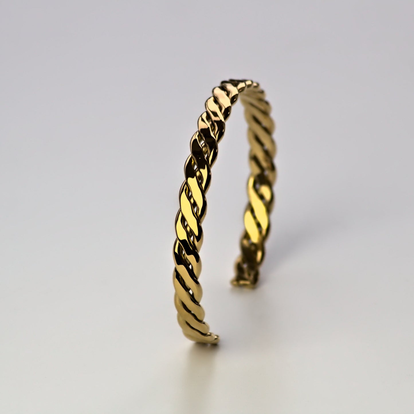 Woven gold cuff bangle, balancing upwards on white flat surface. Light bouncing off the edges, highlighting elegant finish.