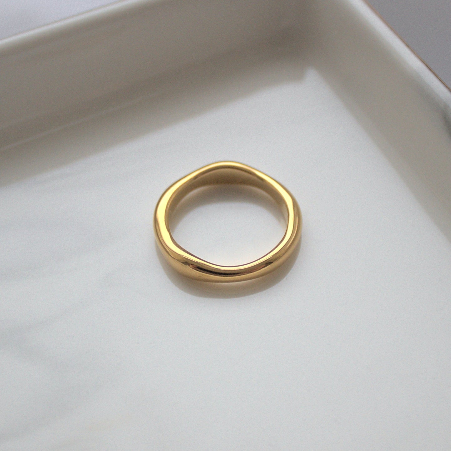 Elegant thicker gold wave ring resting on silver marble tray near corner, light highlighting the smooth ring surface