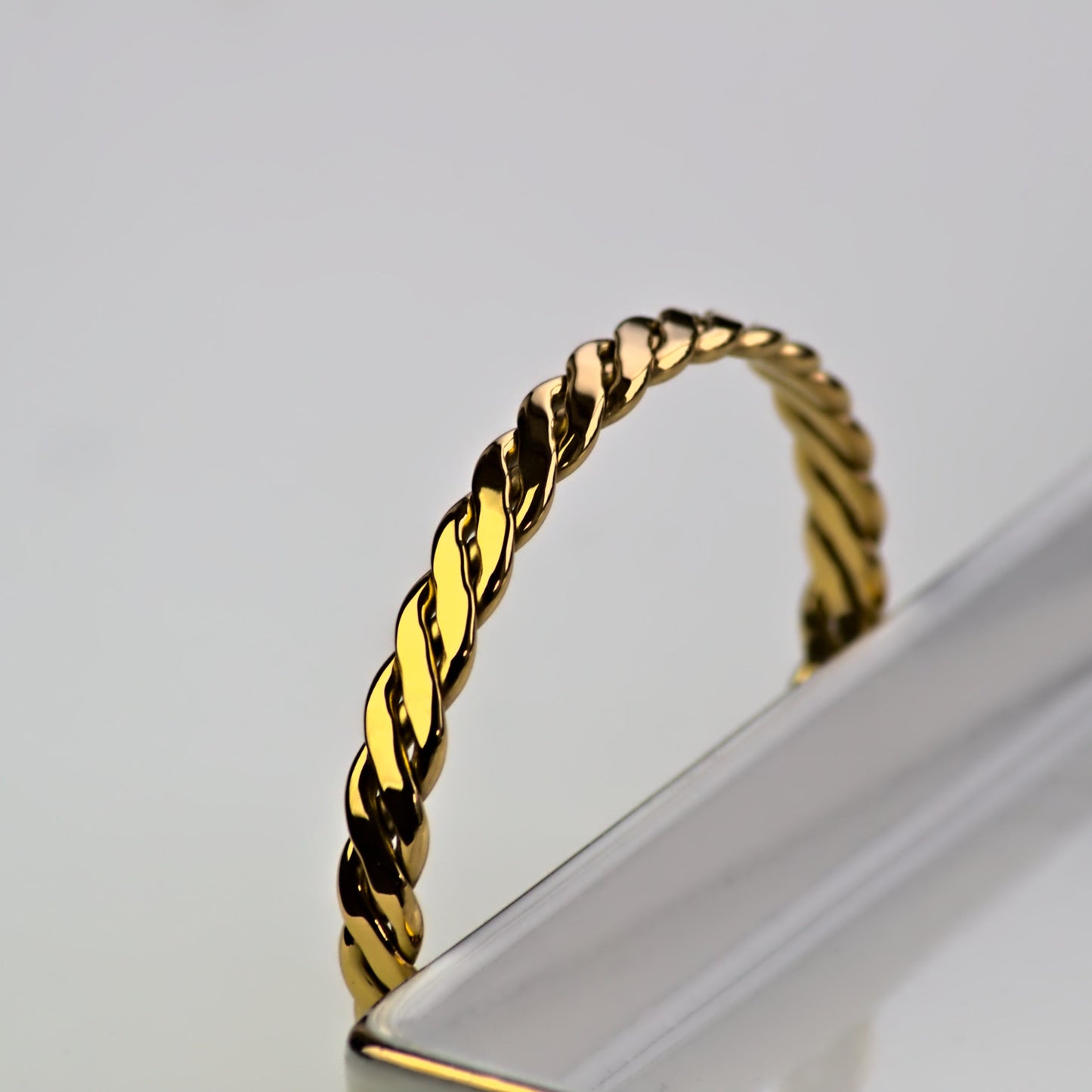 Gold woven bangle, resting against outside edge of marble tray. Only topside visible of bangle, with light shining off of bangle.