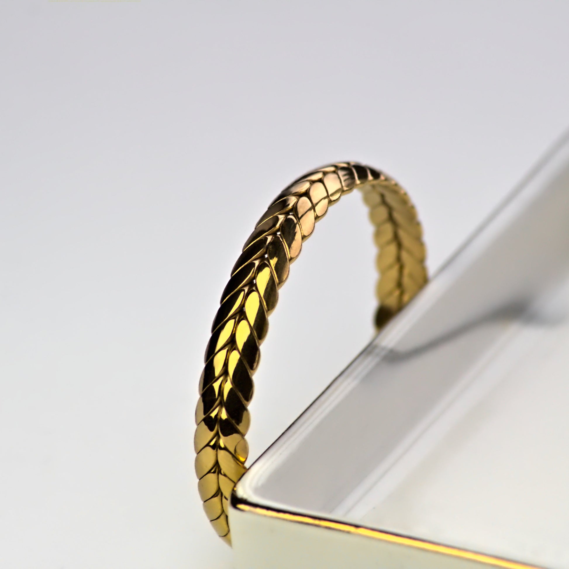Top-side view of ear of wheat bangle resting upright on outwards edge of silver marble tray. Light reflecting on face of bangle.