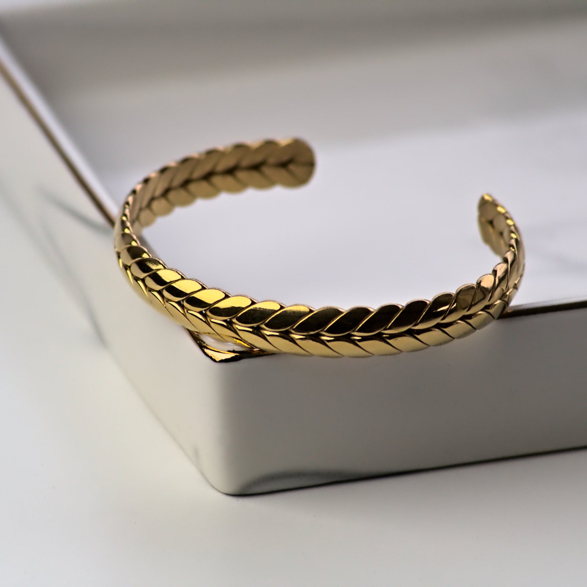 Ear of wheat wheatsheaf gold bangle resting on corner of silver colour marble tray. Front view of bangle with light reflecting off top of bangle.