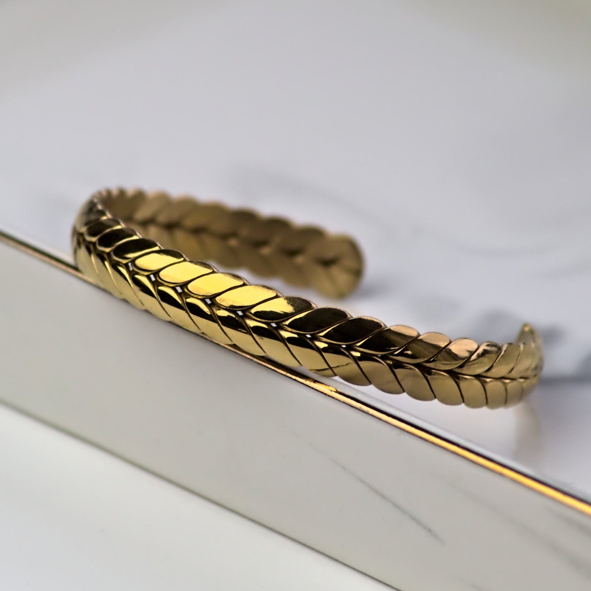 Ear of wheat style gold cuff bangle, resting on edge of silver tray. Close up on front of bangle.