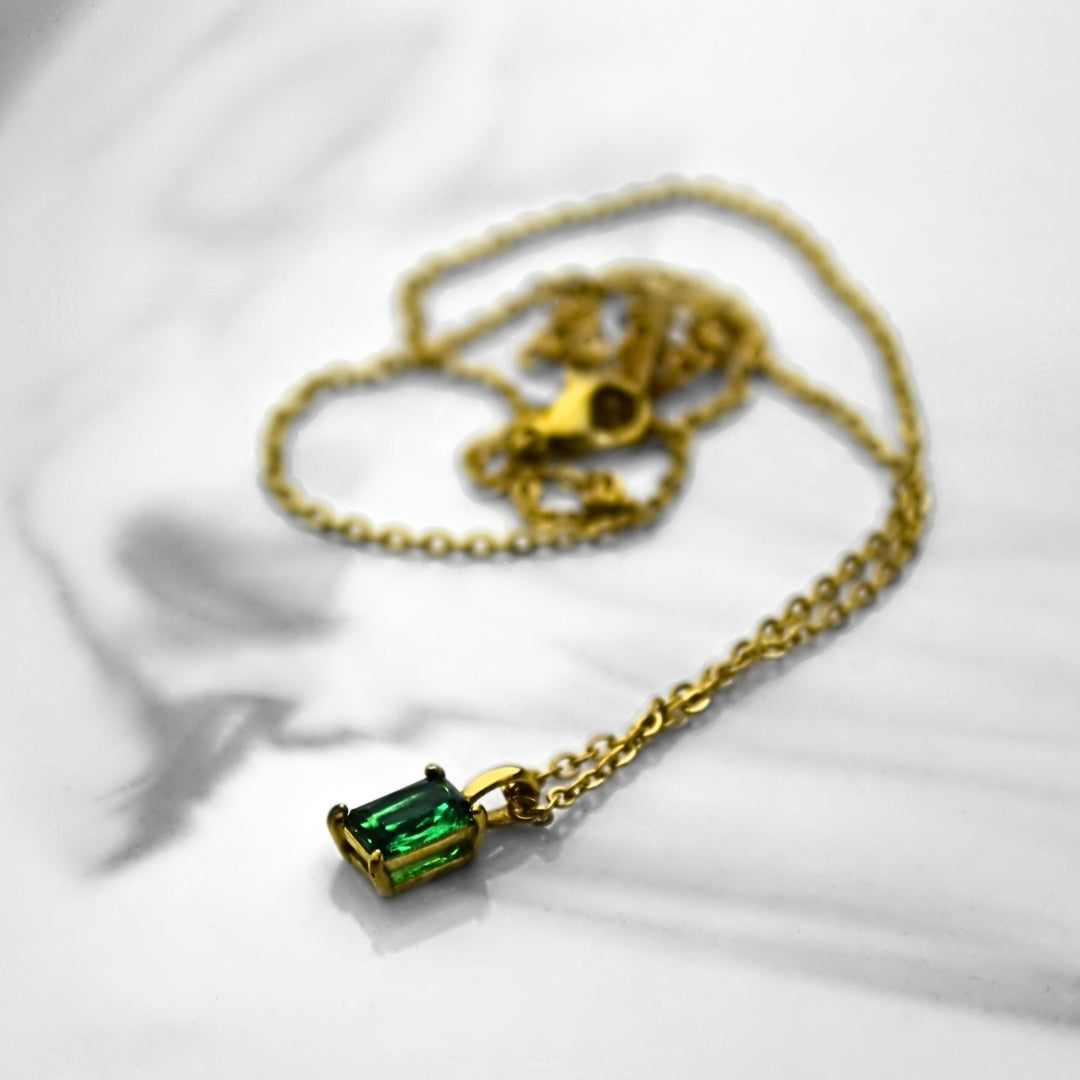 Emerald green pendant necklace and gold chain resting on flat marble surface, with chain resting unorganised in background.