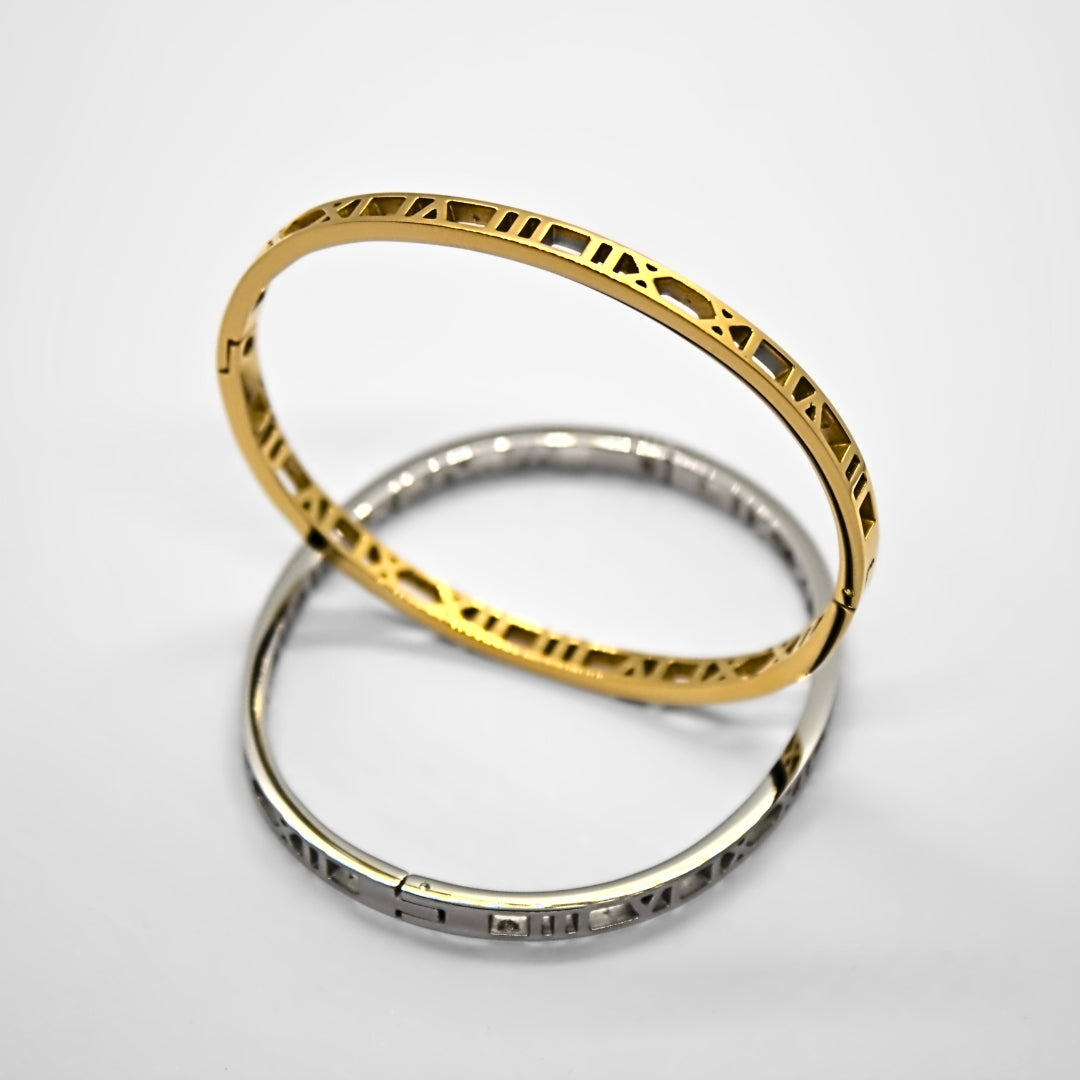 Gold and Silver coloured roman numerals bangle. Silver bangle is resting flat on white surface, gold bangle is balanced upright in the middle of the silver bangle. Perpendicular to each other, with light highlighting the numbers on the top edge of the golden upright bangle. 