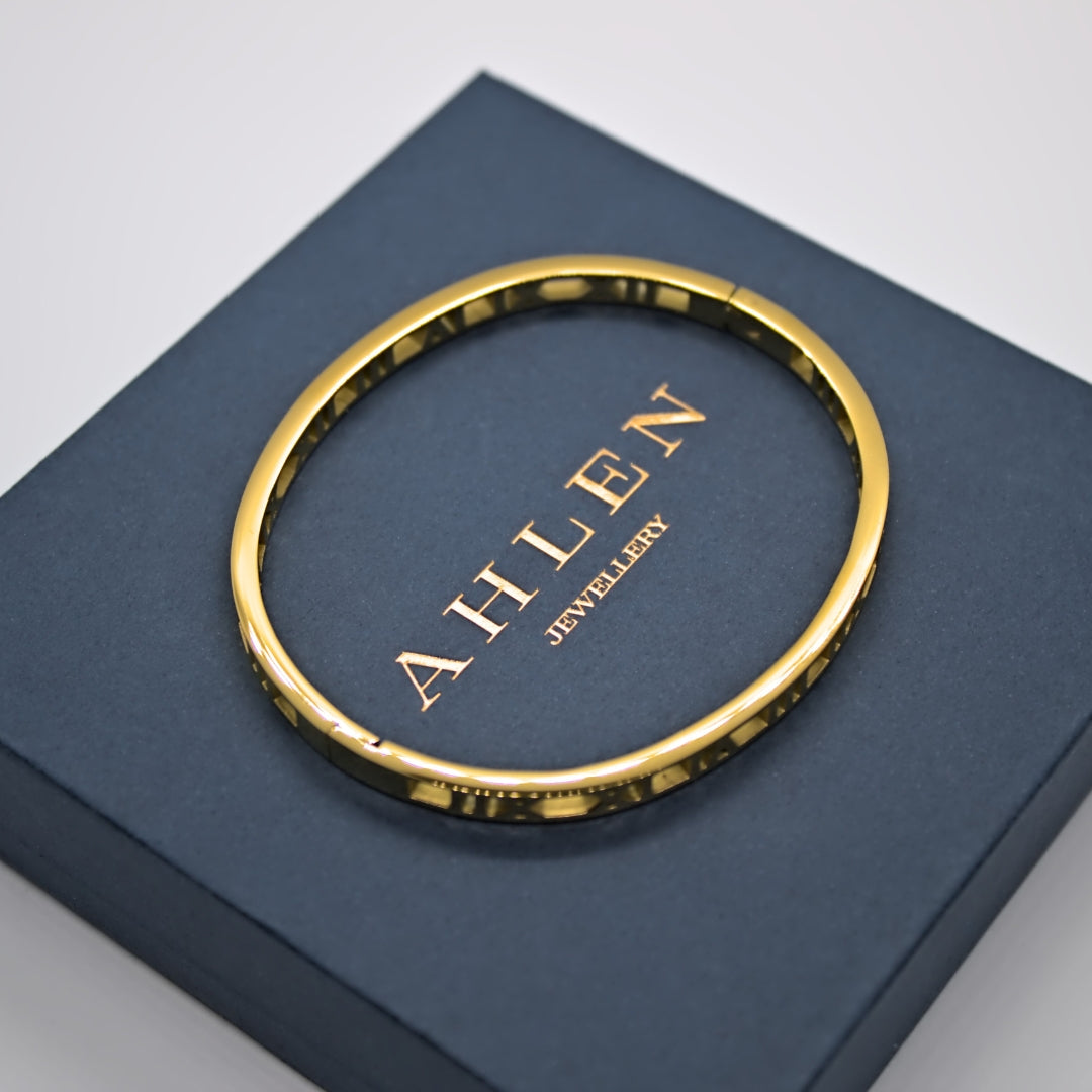 Gold roman numerals bangle resting on blue Ahlen Jewellery gift box, Ahlen Jewellery debossed logo showing in middle of the bangle. Top down view. Box resting on a white background.