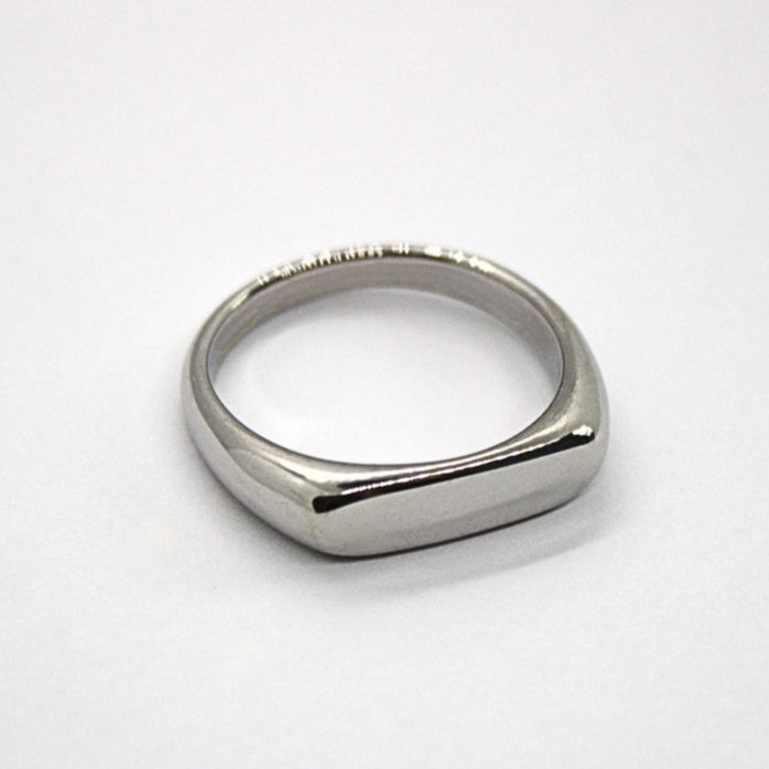 Flat silver signet ring on flat white surface, facing 5 o clock position.